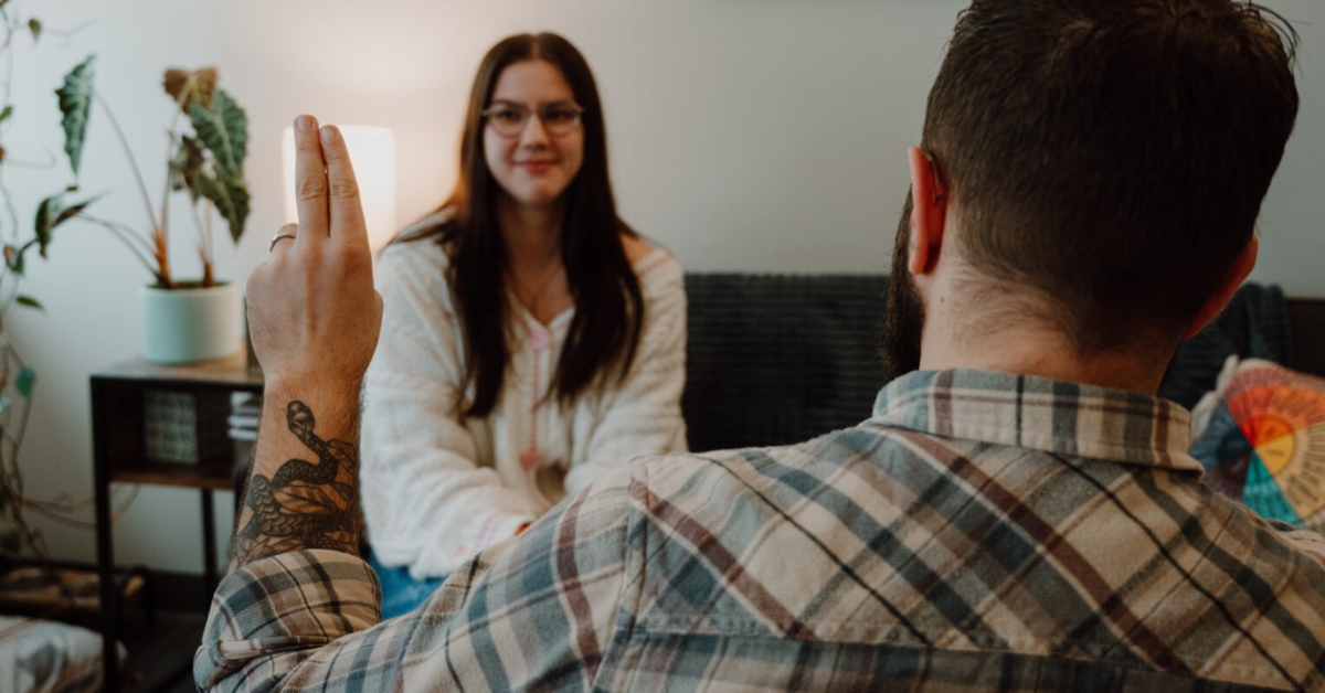 A person with a tattoo raises two fingers while facing another person sitting on a couch, suggesting a conversation or EMDR therapy session in a cozy room—perhaps somewhere in Ogden, Utah.