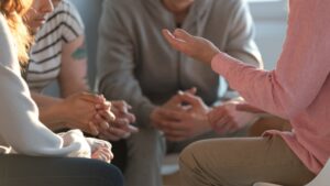 A group of people sit closely together in a circle, with their hands clasped or gesturing as they have a discussion about common questions that often arise in the Family Program.
