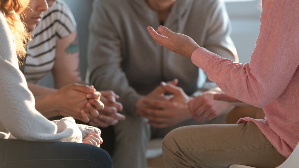 A group of people sit closely together in a circle, with their hands clasped or gesturing as they have a discussion about common questions that often arise in the Family Program.