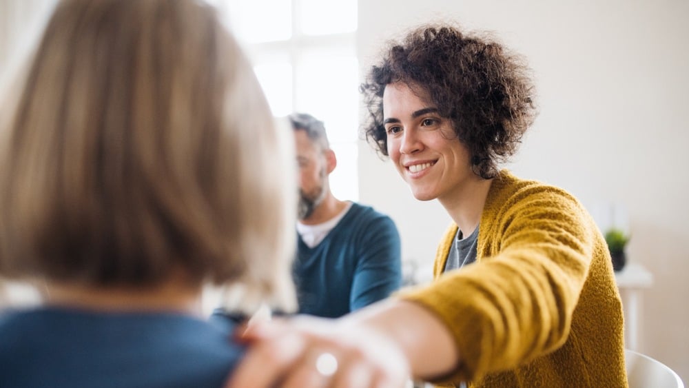 A person with curly hair and a yellow sweater smiles and places a hand on another person’s shoulder during a conversation about addiction recovery in a bright room in Plain City, Utah.