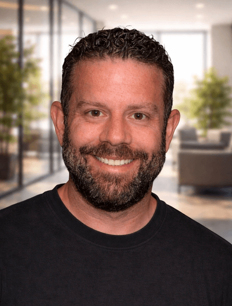 Meet our team: A man with short curly hair and a beard, wearing a dark t-shirt, smiles at the camera in a modern office setting with glass walls and plants.