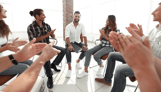 A group of people sit in a circle, smiling and clapping, in a bright room with large windows and a brick pillar at a local addiction treatment center.