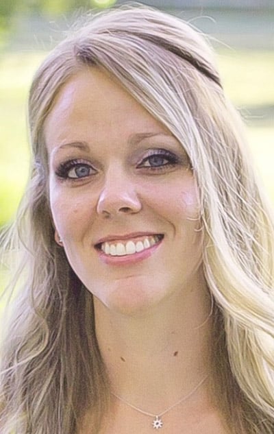Meet our team: A woman with long blonde hair and blue eyes smiles at the camera, wearing a necklace with a star-shaped pendant. The background is blurred with greenery.