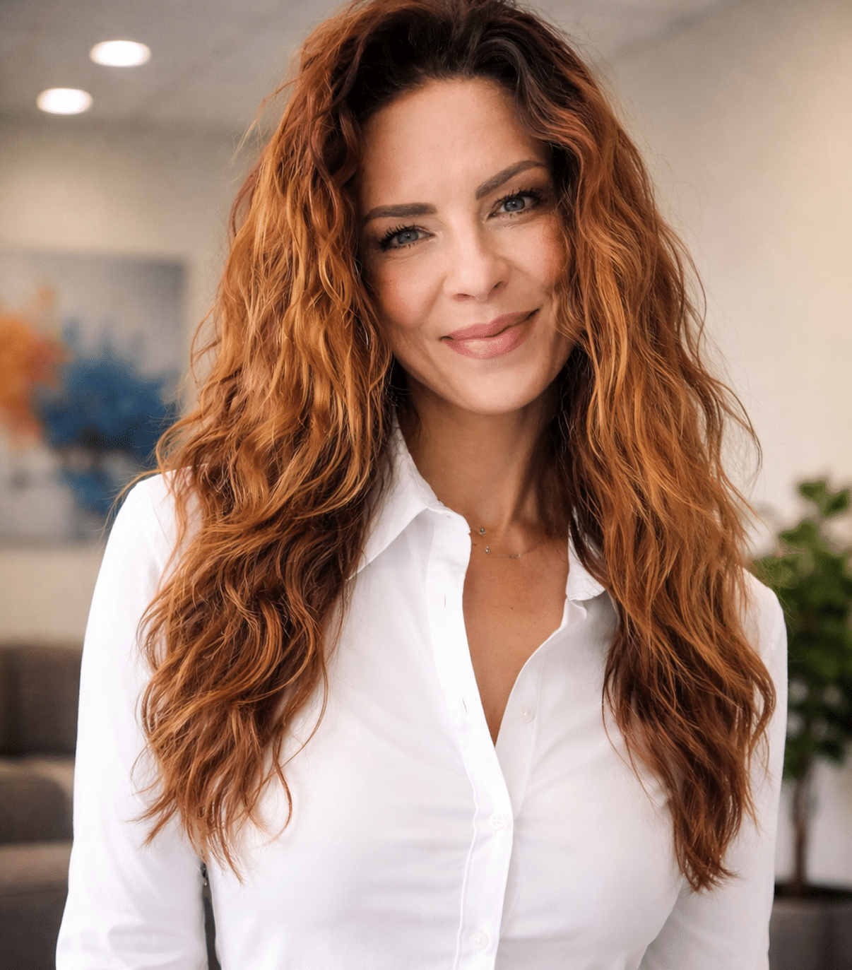 Meet our team: A woman with long wavy red hair, wearing a white button-up shirt, stands indoors with a slight smile.