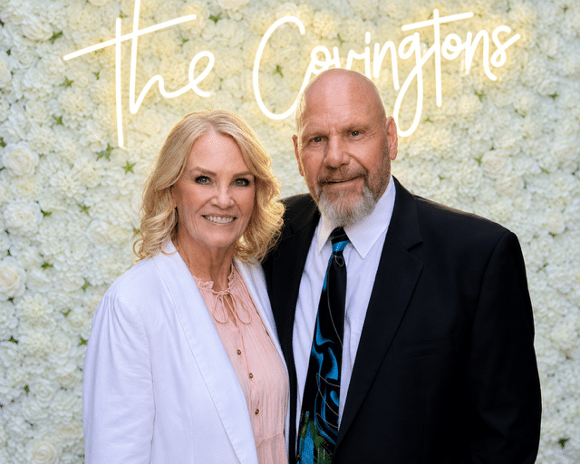 A woman and a man in formal attire stand together, smiling, in front of a white flower wall with “The Covingtons” written in the background—an elegant snapshot that invites you to meet our team.