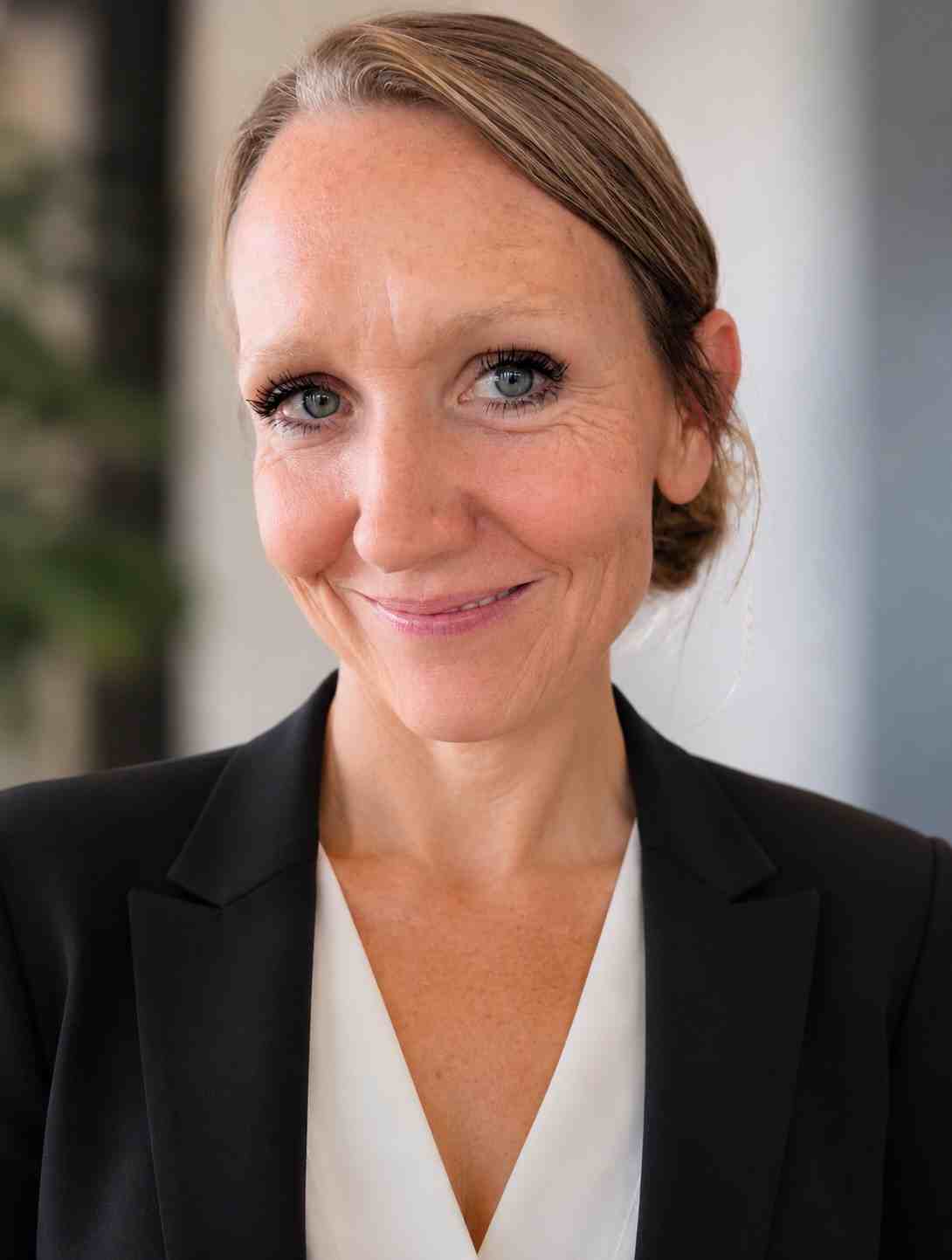 Meet our team: A woman with light skin and blue eyes, wearing a black blazer and white top, smiles at the camera in a professional indoor setting.
