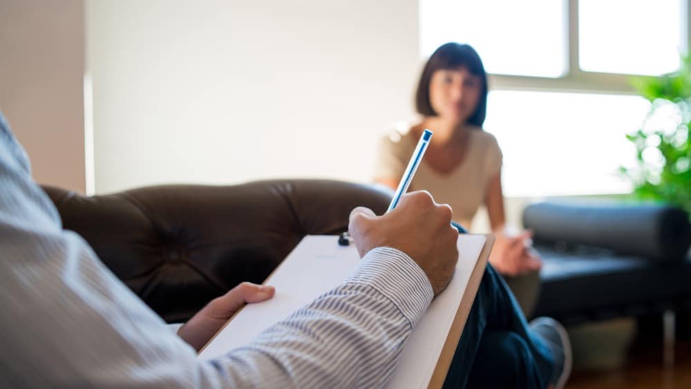 Person holding clipboard and pen, taking notes while sitting across from another person in a well-lit room—a supportive setting often found at Recovery Services in Farr West UT, dedicated to guiding individuals on their journey to addiction recovery.