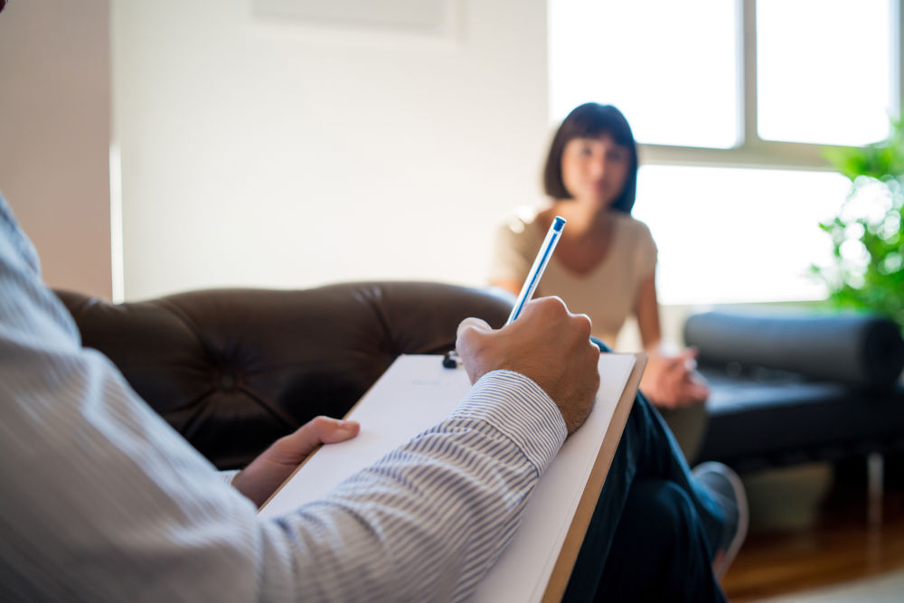 A person holding a clipboard writes notes while listening to another person sitting on a couch in a well-lit room, illustrating a typical Drug Addiction Treatment session.