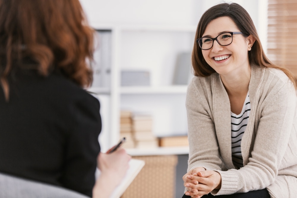 A woman wearing glasses and a beige sweater smiles while talking to another person, who is holding a notepad and pen in a bright office setting focused on addiction recovery services in Farr West, UT.
